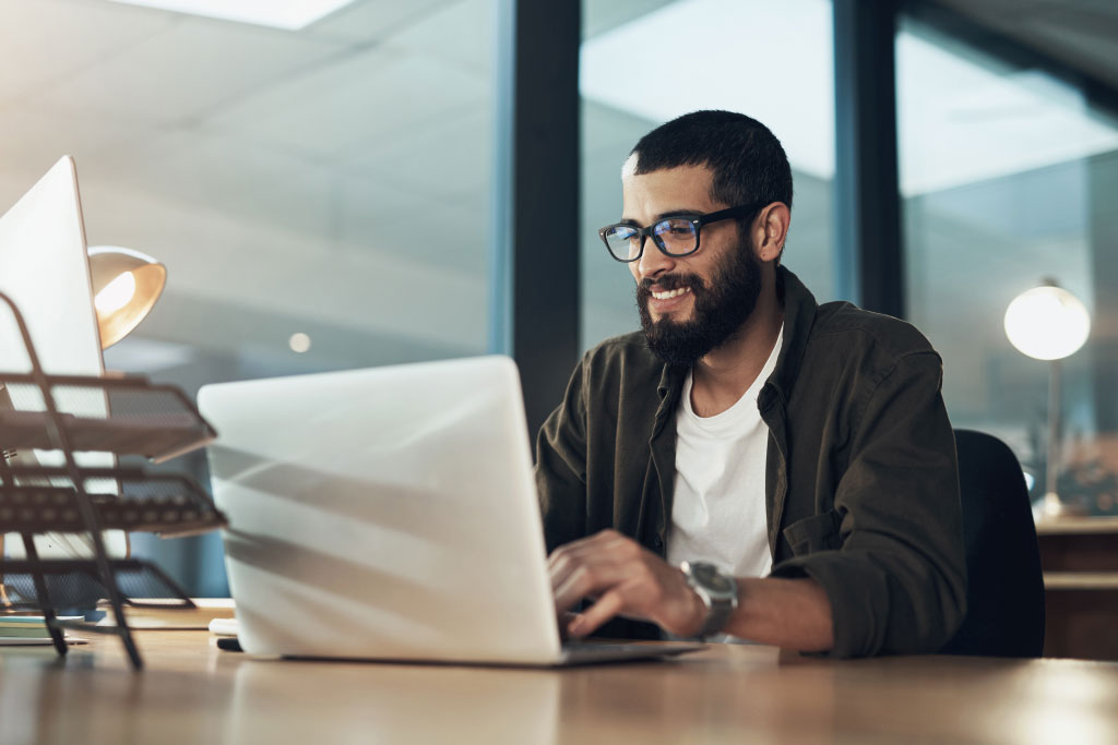 A Young Man Smiling While Working On A Laptop