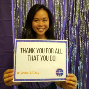 A woman holds a paper sign that says, "Thank you for all that you do," for Global TA Day