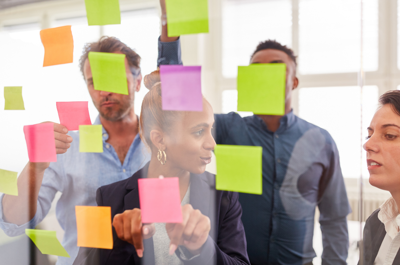 A group of workers standing in front of a glass wall of post-it notes to represent people analytics